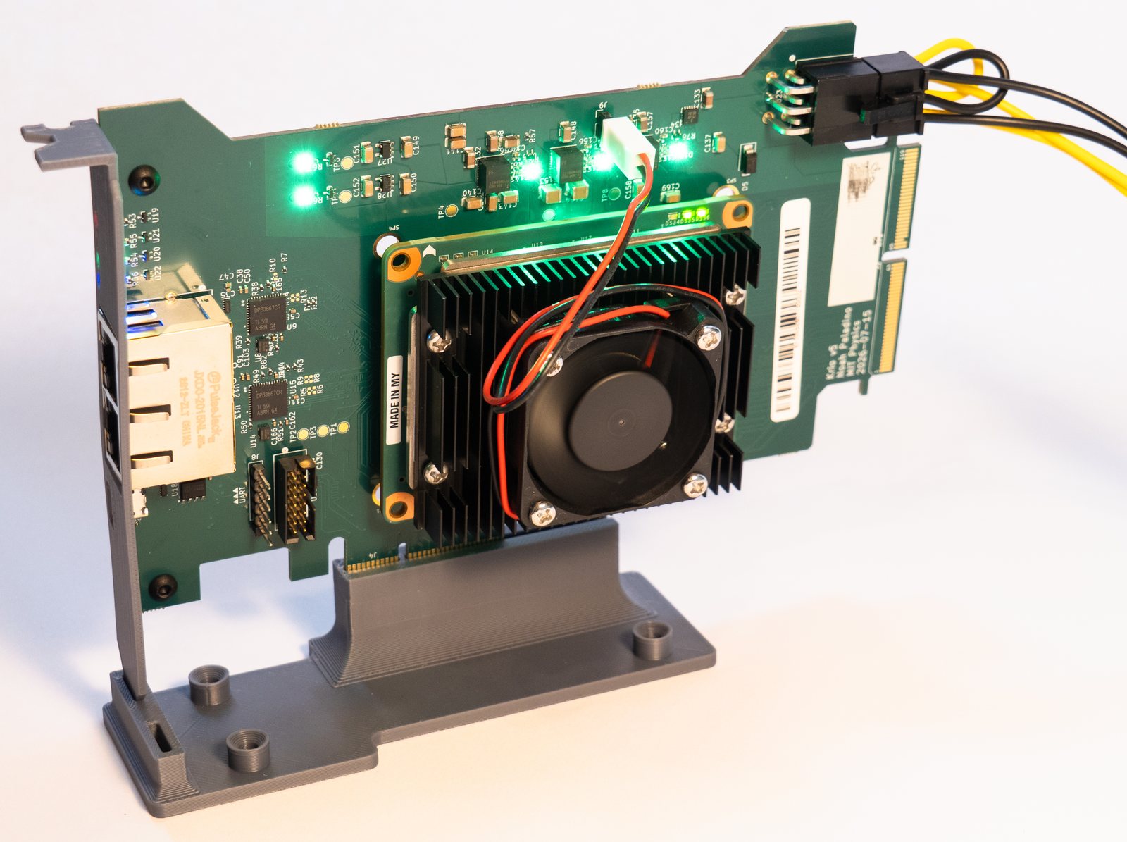 The KrIO carrier stood upright in a 3D-printed bench stand, seen from the front, green power LEDs lit across the top edge and the Kria K26's heatsink and fan facing the camera.